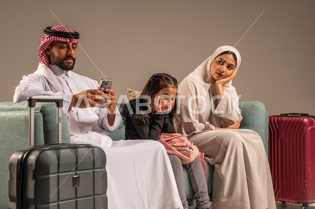 انتظار موعد الطائرة بايماءات الملل، بورتريه لرجل عربي خليجي سعودي يرتدي الثوب التقليدي والشماغ يجلس على كرسي مريح مع زوجته وابنته يمسك حقيبة الأمتعة، الإستعداد لقضاء الإجازة الصيفية والذهاب في رحلة سفر ممتعة، الأجواء العائلية المليئة بالحب،  خلفية رمادية