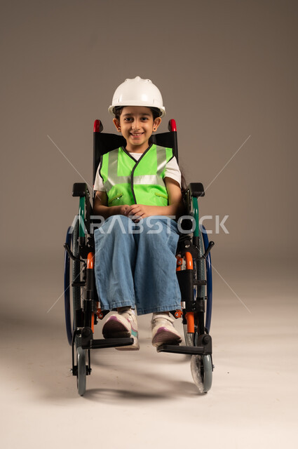 دمج الأطفال ذوي الهمم في المجتمع، النظر إلى الكاميرا بإيماءات السعادة والسرور، بورتريه لولد من ذوي الاحتياجات الخاصة عربي خليجي سعودي يرتدي سترة وخوذة الحماية يجلس على كرسي متحرك، الإعاقة الحركية وعدم القدرة على المشي، الجسم بالطول الكامل، خلفية بيج