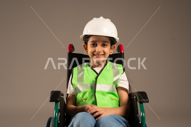 دمج الأطفال ذوي الهمم في المجتمع، النظر إلى الكاميرا بإيماءات السعادة والسرور، بورتريه مقرب لولد من ذوي الاحتياجات الخاصة عربي خليجي سعودي يرتدي سترة وخوذة الحماية يجلس على كرسي متحرك، الإعاقة الحركية وعدم القدرة على المشي، خلفية بيج