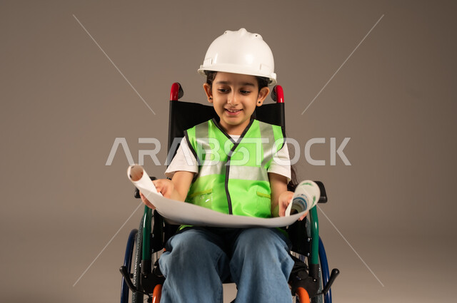 الإعاقة الحركية وعدم القدرة على المشي، الإمساك بمخطط الإعمار وتفحصه، بورتريه مقرب لولد من ذوي الاحتياجات الخاصة عربي خليجي سعودي يرتدي سترة وخوذة الحماية يجلس على كرسي متحرك يستمتع بتجريب مهنة المهندس منذ الصغر، دمج اطفال ذوي الهمم في المجتمع، خلفية بيج