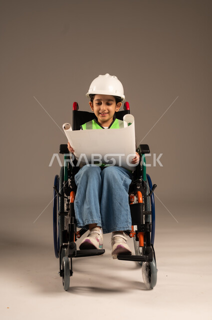 الإمساك بمخطط الإعمار وتفحصه، بورتريه لولد من ذوي الاحتياجات الخاصة عربي خليجي سعودي يرتدي سترة وخوذة الحماية يجلس على كرسي متحرك يجرب مهنة المهندس منذ الصغر، الإعاقة الحركية وعدم القدرة على المشي، دمج اطفال ذوي الهمم في المجتمع، الجسم بالطول الكامل، خلفية بيج