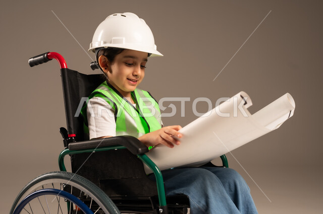 الإعاقة الحركية وعدم القدرة على المشي، الإمساك بمخطط الإعمار وتفحصه، بورتريه مقرب لولد من ذوي الاحتياجات الخاصة عربي خليجي سعودي يرتدي سترة وخوذة الحماية يجلس على كرسي متحرك يستمتع بتجريب مهنة المهندس منذ الصغر، دمج اطفال ذوي الهمم في المجتمع، خلفية بيج