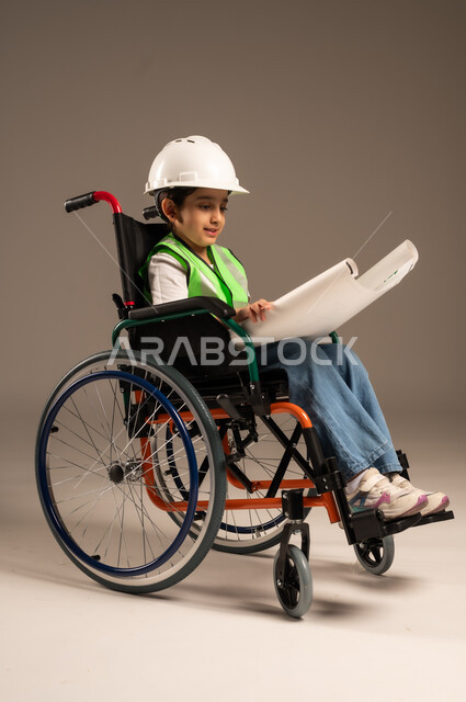 الإمساك بمخطط الإعمار وتفحصه، بورتريه لولد من ذوي الاحتياجات الخاصة عربي خليجي سعودي يرتدي سترة وخوذة الحماية يجلس على كرسي متحرك يجرب مهنة المهندس منذ الصغر، الإعاقة الحركية وعدم القدرة على المشي، دمج اطفال ذوي الهمم في المجتمع، الجسم بالطول الكامل، خلفية بيج