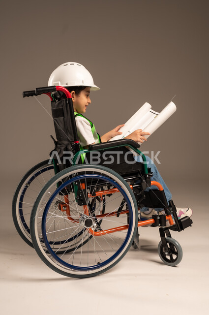 الإمساك بمخطط الإعمار وتفحصه، بورتريه لولد من ذوي الاحتياجات الخاصة عربي خليجي سعودي يرتدي سترة وخوذة الحماية يجلس على كرسي متحرك يجرب مهنة المهندس منذ الصغر، الإعاقة الحركية وعدم القدرة على المشي، دمج اطفال ذوي الهمم في المجتمع، الجسم بالطول الكامل، خلفية بيج