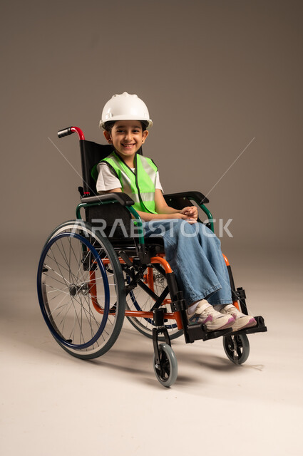 الإعاقة الحركية وعدم القدرة على المشي، الإمساك بمخطط الإعمار وتفحصه، بورتريه مقرب لولد من ذوي الاحتياجات الخاصة عربي خليجي سعودي يرتدي سترة وخوذة الحماية يجلس على كرسي متحرك يستمتع بتجريب مهنة المهندس منذ الصغر، دمج اطفال ذوي الهمم في المجتمع، خلفية بيج