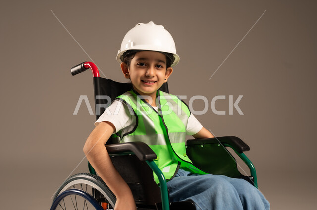 النظر إلى الكاميرا بإيماءات السعادة والسرور، بورتريه مقرب لولد من ذوي الاحتياجات الخاصة عربي خليجي سعودي يرتدي سترة وخوذة الحماية يجلس على كرسي متحرك، الإعاقة الحركية وعدم القدرة على المشي، دمج اطفال ذوي الهمم في المجتمع، خلفية بيج