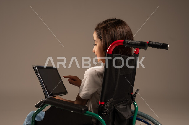 استخدام الأجهزة والتكنولوجيا الحديثة، التسلية واللعب الالكتروني، بورتريه لفتاة عربية خليجية سعودية من ذوي الاحتياجات الخاصة ترتدي الزي الكاجوال تجلس على كرسي متحرك تمسك بيدها الجهاز اللوحي، تحسين حياة ذوي الإعاقة الحركية، خلفية رمادية