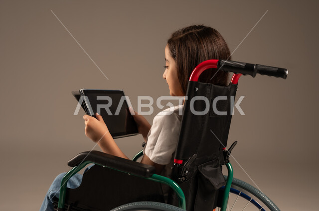 استخدام الأجهزة والتكنولوجيا الحديثة، التسلية واللعب الالكتروني، بورتريه لفتاة عربية خليجية سعودية من ذوي الاحتياجات الخاصة ترتدي الزي الكاجوال تجلس على كرسي متحرك تمسك بيدها الجهاز اللوحي، تحسين حياة ذوي الإعاقة الحركية، خلفية رمادية