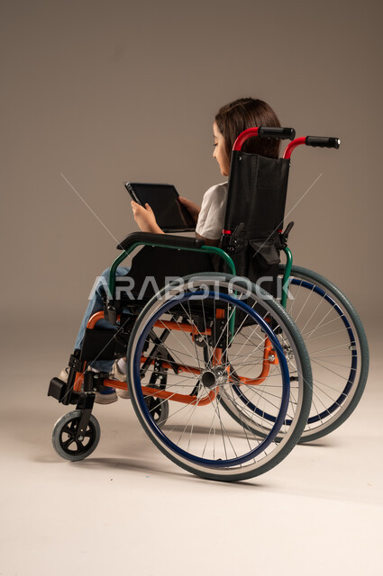 استخدام الأجهزة والتكنولوجيا الحديثة، التسلية واللعب الالكتروني، بورتريه لفتاة عربية خليجية سعودية من ذوي الاحتياجات الخاصة ترتدي الزي الكاجوال تجلس على كرسي متحرك تمسك بيدها الجهاز اللوحي، تحسين حياة ذوي الإعاقة الحركية، خلفية رمادية