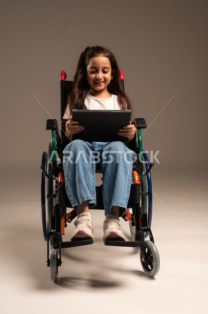 التسلية واللعب الالكتروني، استخدام الأجهزة والتكنولوجيا الحديثة، بورتريه لفتاة عربية خليجية سعودية من ذوي الاحتياجات الخاصة ترتدي الزي الكاجوال تجلس على كرسي متحرك تمسك بيدها الجهاز اللوحي، تحسين حياة ذوي الإعاقة الحركية، خلفية رمادية