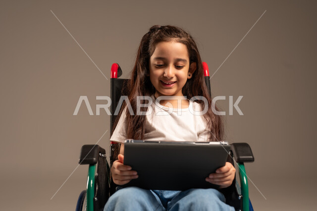 التسلية واللعب الالكتروني، استخدام الأجهزة والتكنولوجيا الحديثة، بورتريه لفتاة عربية خليجية سعودية من ذوي الاحتياجات الخاصة ترتدي الزي الكاجوال تجلس على كرسي متحرك تمسك بيدها الجهاز اللوحي، تحسين حياة ذوي الإعاقة الحركية، خلفية رمادية