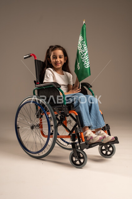 مشاركة ذوي الهمم بالأعياد والمناسبات الوطنية, استذكار اليوم الوطني السعودي في23 سبتمبر,  بورتريه لفتاة عربية خليجية سعودية ترتدي الزي الكاجوال تجلس على كرسي متحرك تحمل علم السعودية باحترام وفخر، التعبير عن حب الوطن والانتماء إليه, خلفية رمادية