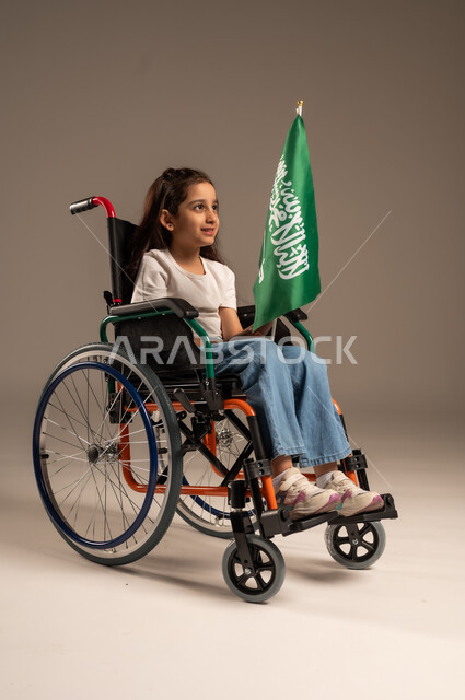 Participation of people with disabilities in national holidays and occasions, remembering Saudi National Day on September 23, portrait of a Saudi Gulf Arab girl wearing casual clothes sitting in a wheelchair holding the Saudi flag with respect and pride, expressing love for the homeland and belonging to it, gray background