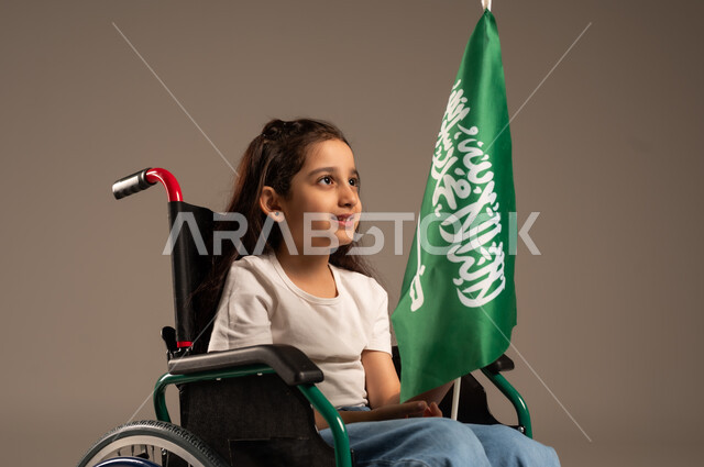 مشاركة ذوي الهمم بالأعياد والمناسبات الوطنية, استذكار اليوم الوطني السعودي في23 سبتمبر,  بورتريه لفتاة عربية خليجية سعودية ترتدي الزي الكاجوال تجلس على كرسي متحرك تحمل علم السعودية باحترام وفخر، التعبير عن حب الوطن والانتماء إليه, خلفية رمادية