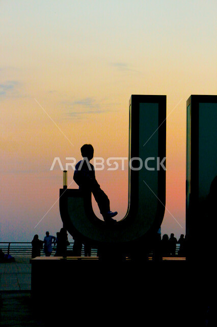 طفل يجلس على شعار جدة في كورنيش جدة الواجهة البحرية السياحية في المملكة العربية السعودية , غروب الشمس على كورنيش جدة ، منظر الغروب على الشاطئ ، ​مناظر طبيعية خلابة في المملكة العربية السعودية