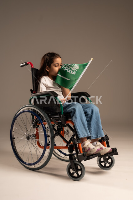 Participation of people with disabilities in national holidays and occasions, remembering Saudi National Day on September 23, portrait of a Saudi Gulf Arab girl wearing casual clothes sitting in a wheelchair holding the Saudi flag with respect and pride, expressing love for the homeland and belonging to it, gray background