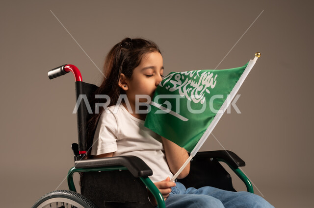 Participation of people with disabilities in national holidays and occasions, remembering Saudi National Day on September 23, portrait of a Saudi Gulf Arab girl wearing casual clothes sitting in a wheelchair holding the Saudi flag with respect and pride, expressing love for the homeland and belonging to it, gray background