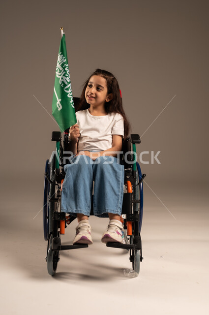 استذكار اليوم الوطني السعودي في23 سبتمبر,  بورتريه لفتاة عربية خليجية سعودية ترتدي الزي الكاجوال تجلس على كرسي متحرك تحمل علم السعودية باحترام وفخر، مشاركة ذوي الهمم بالأعياد والمناسبات الوطنية, التعبير عن حب الوطن والانتماء إليه, خلفية رمادية