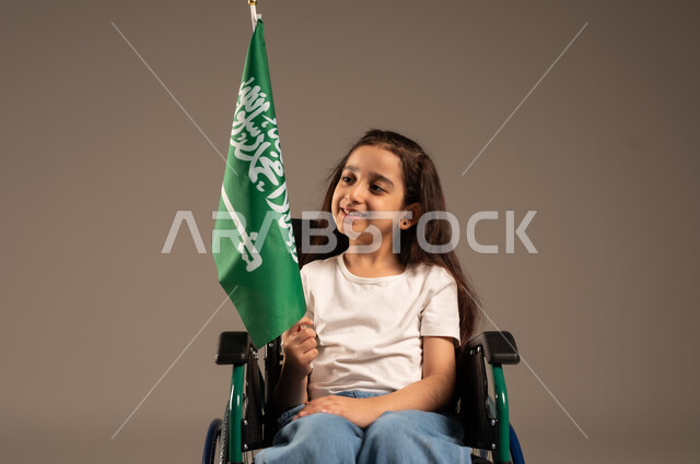 استذكار اليوم الوطني السعودي في23 سبتمبر,  بورتريه لفتاة عربية خليجية سعودية ترتدي الزي الكاجوال تجلس على كرسي متحرك تحمل علم السعودية باحترام وفخر، مشاركة ذوي الهمم بالأعياد والمناسبات الوطنية, التعبير عن حب الوطن والانتماء إليه, خلفية رمادية