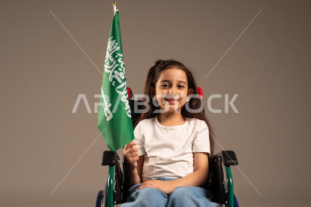 استذكار اليوم الوطني السعودي في23 سبتمبر,  بورتريه لفتاة عربية خليجية سعودية ترتدي الزي الكاجوال تجلس على كرسي متحرك تحمل علم السعودية باحترام وفخر، مشاركة ذوي الهمم بالأعياد والمناسبات الوطنية, التعبير عن حب الوطن والانتماء إليه, خلفية رمادية