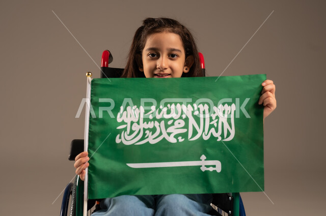 استذكار اليوم الوطني السعودي في23 سبتمبر,  بورتريه لفتاة عربية خليجية سعودية ترتدي الزي الكاجوال تجلس على كرسي متحرك تحمل علم السعودية باحترام وفخر، مشاركة ذوي الهمم بالأعياد والمناسبات الوطنية, التعبير عن حب الوطن والانتماء إليه, خلفية رمادية