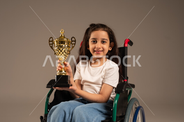 التتويج بالبطولة بعد الفوز والحصول على المركز الأول، بورتريه لفتاة عربية خليجية سعودية ترتدي الزي الكاجوال تجلس على كرسي متحرك تحمل بيدها الكأس الذهبي بحماس، تحسين حياة ذوي الإعاقة الحركية، مفهوم الربح والفوز، خلفية رمادية