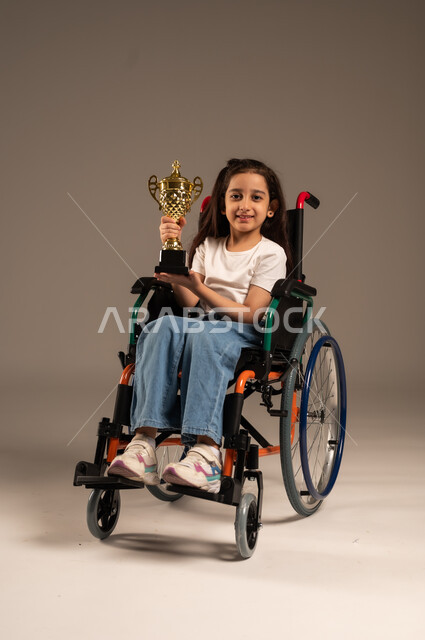 التتويج بالبطولة بعد الفوز والحصول على المركز الأول، بورتريه لفتاة عربية خليجية سعودية ترتدي الزي الكاجوال تجلس على كرسي متحرك تحمل بيدها الكأس الذهبي بحماس، تحسين حياة ذوي الإعاقة الحركية، مفهوم الربح والفوز، خلفية رمادية