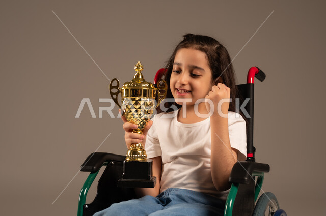 Crowning the championship after winning and getting first place, portrait of a Saudi Arabian Gulf girl wearing casual clothes sitting on a wheelchair holding the golden cup in her hand with enthusiasm, improving the lives of people with motor disabilities, the concept of profit and victory, gray background