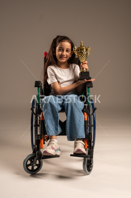 التتويج بالبطولة بعد الفوز والحصول على المركز الأول، بورتريه لفتاة عربية خليجية سعودية ترتدي الزي الكاجوال تجلس على كرسي متحرك تحمل بيدها الكأس الذهبي بحماس، تحسين حياة ذوي الإعاقة الحركية، مفهوم الربح والفوز، خلفية رمادية