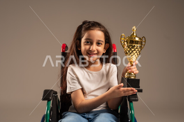 Crowning the championship after winning and getting first place, portrait of a Saudi Arabian Gulf girl wearing casual clothes sitting on a wheelchair holding the golden cup in her hand with enthusiasm, improving the lives of people with motor disabilities, the concept of profit and victory, gray background