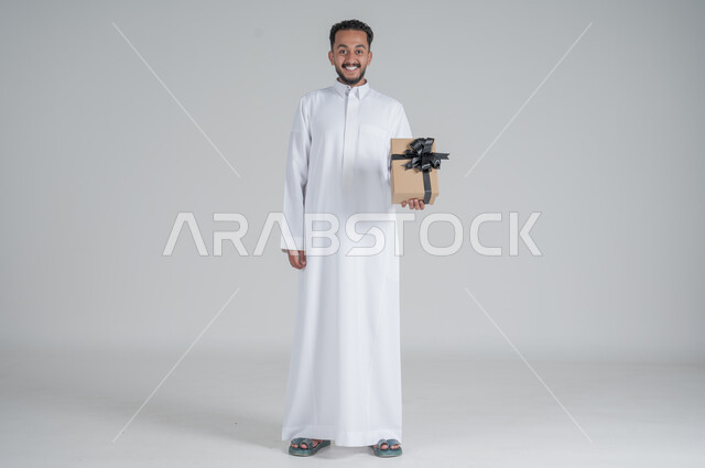 مفهوم الشكر والامتنان، تقديم الهدايا بالاعياد والمناسبات الخاصة، التعبير عن الحب والتقدير والاحترام، بورتريه لرجل عربي سعودي يرتدي الثوب التقليدي يحمل بيديه صندوق هدية أسود, الجسم بالطول الكامل، خلفية رمادي