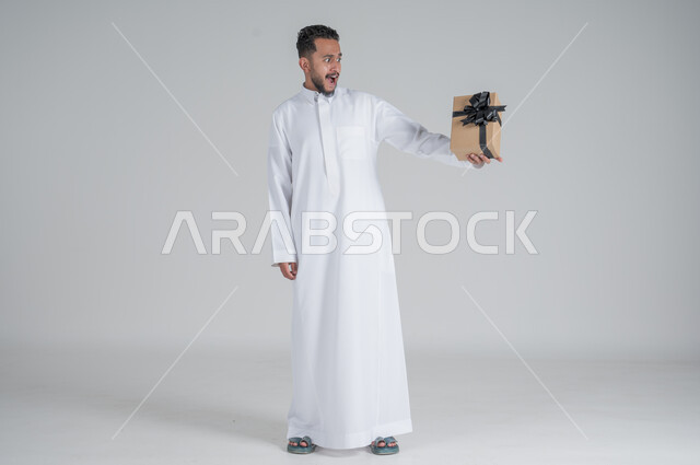 The concept of thanks and gratitude, giving gifts on holidays and special occasions, expressing love, appreciation and respect, portrait of a Saudi Arab man wearing a traditional thobe holding a black gift box in his hands, full-length body, gray background