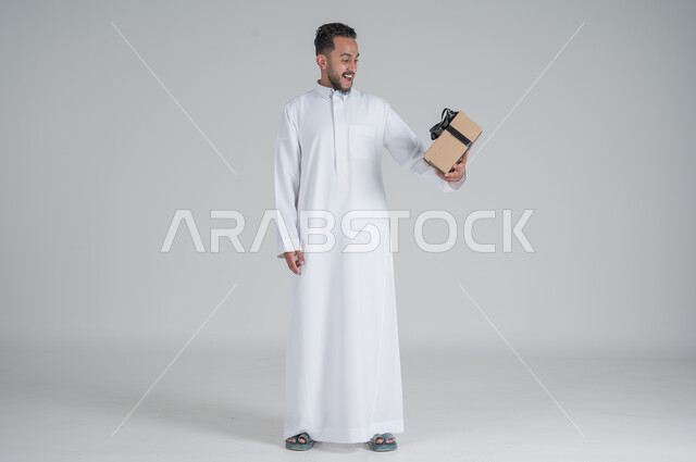 مفهوم الشكر والامتنان، تقديم الهدايا بالاعياد والمناسبات الخاصة، التعبير عن الحب والتقدير والاحترام، بورتريه لرجل عربي سعودي يرتدي الثوب التقليدي يحمل بيديه صندوق هدية أسود, الجسم بالطول الكامل، خلفية رمادي