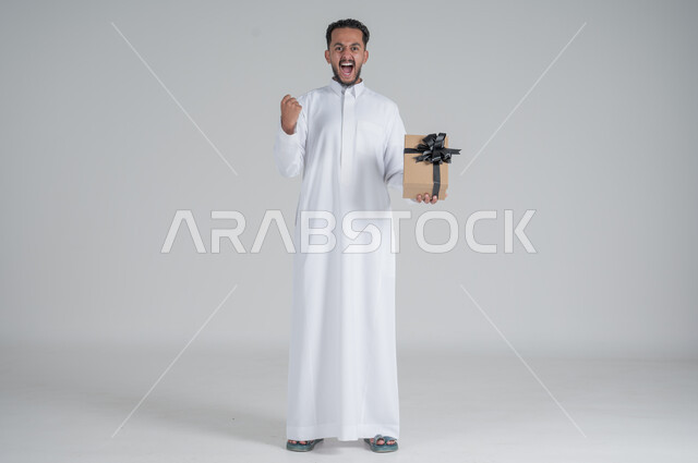 مفهوم الشكر والامتنان، تقديم الهدايا بالاعياد والمناسبات الخاصة، التعبير عن الحب والتقدير والاحترام، بورتريه لرجل عربي سعودي يرتدي الثوب التقليدي يحمل بيديه صندوق هدية أسود, الجسم بالطول الكامل، خلفية رمادي