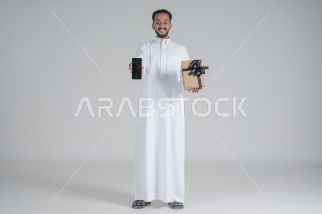 مفهوم الشكر والامتنان، تقديم الهدايا بالاعياد والمناسبات الخاصة، التعبير عن الحب والتقدير والاحترام، بورتريه لرجل عربي سعودي يرتدي الثوب التقليدي يحمل بيد علبة هدية وباليد الاخرى هاتف محمول بشاشة سوداء, الجسم بالطول الكامل، خلفية رمادي