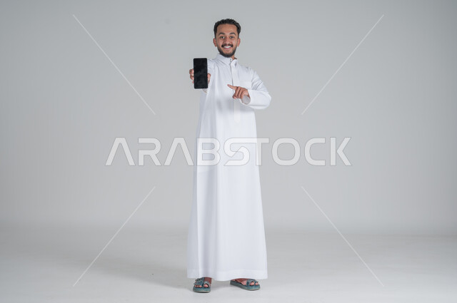 مفهوم التسويق الرقمي الالكتروني، عرض تطبيقات وخدمات الهاتف المحمول، بورتريه لشاب عربي خليجي سعودي مبتسم يرتدي الثوب التقليدي يحمل بيده جوال بشاشة سوداء فارغة ويشير اليه، استخدام التكنولوجيا المتطورة، صورة الجسم بالطول الكامل، خلفية رمادي