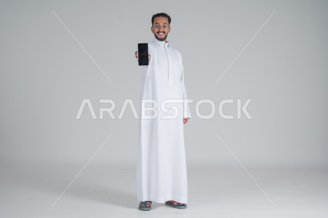 مفهوم التسويق الرقمي الالكتروني، عرض تطبيقات وخدمات الهاتف المحمول، بورتريه لشاب عربي خليجي سعودي مبتسم يرتدي الثوب التقليدي يحمل بيده جوال بشاشة سوداء فارغة، استخدام التكنولوجيا المتطورة، صورة الجسم بالطول الكامل، خلفية رمادي