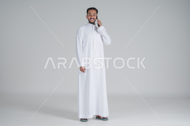 Following the latest developments in the world, using modern techniques and technology, a portrait of a young Saudi Arabian Gulf man wearing a traditional dress, communicating with family and relatives via mobile phone, chatting with friends, full-length body, gray background
