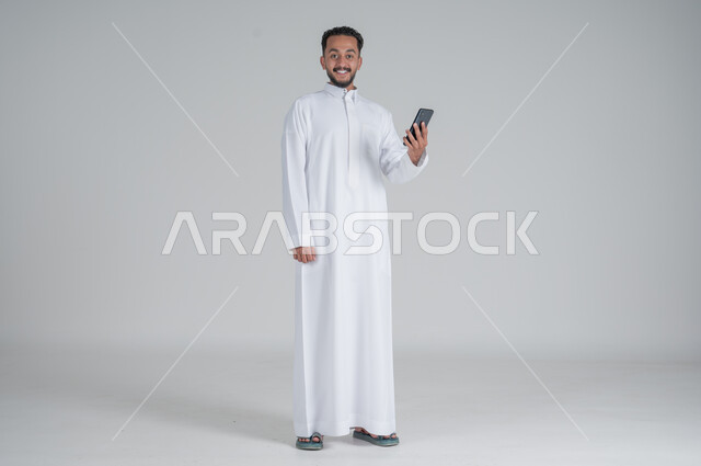 متابعة اّخر مستجدات العالم، استخدام التقنيات الحديثة والتكنولوجيا، بورتريه لشاب عربي سعودي خليجي يرتدي الثوب التقليدي يتواصل مع الاهل والاقارب عبر الهاتف المحمول، الدردشة مع الاصدقاء, الجسم بالطول الكامل، خلفية رمادي
