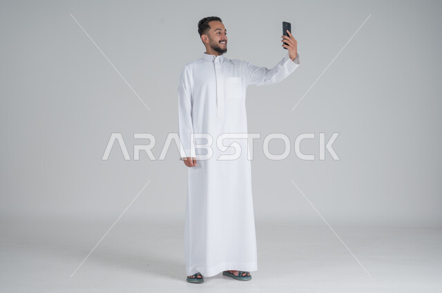 متابعة اّخر مستجدات العالم، استخدام التقنيات الحديثة والتكنولوجيا، بورتريه لشاب عربي سعودي خليجي يرتدي الثوب التقليدي يتواصل مع الاهل والاقارب عبر الهاتف المحمول، الدردشة مع الاصدقاء, الجسم بالطول الكامل، خلفية رمادي