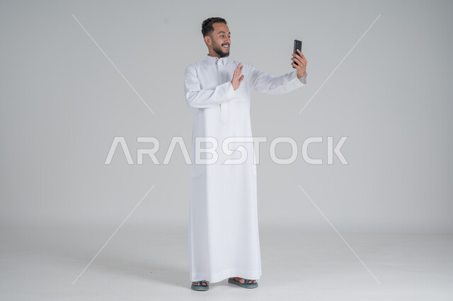 Following the latest developments in the world, using modern techniques and technology, a portrait of a young Saudi Arabian Gulf man wearing a traditional dress, communicating with family and relatives via mobile phone, chatting with friends, full-length body, gray background