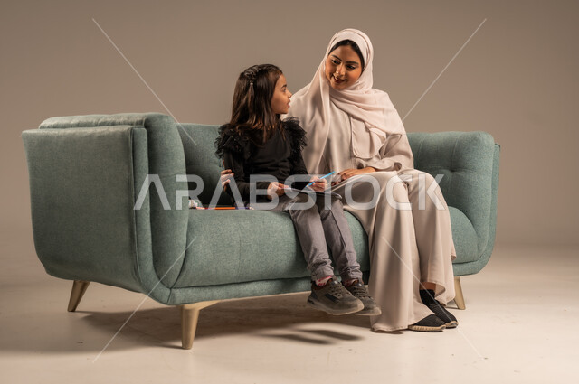 الاهتمام بتعليم الأبناء ودعمهم، بورتريه لام عربية سعودية خليجية ترتدي عباءة ملونة تجلس على كرسي مريح تقوم بتدريس ابنتها، المساعدة في تأدية الواجبات المدرسية، الانشطة التعليمية وتنمية القدرات، خلفية رمادية