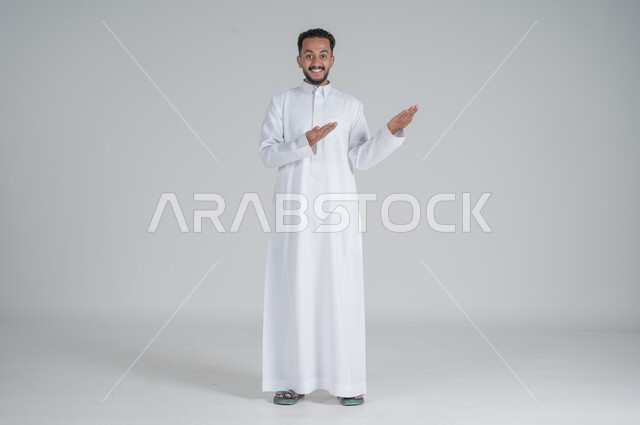 الوقوف باستقامة واعتدال, الإشارة إلى شيء ما, بورتريه لرجل عربي خليجي سعودي يرتدي الثوب التقليدي يشير بيديه إلى شيء ما, صورة الجسم بالطول الكامل, خلفية رمادي