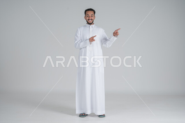 الوقوف باستقامة واعتدال, الإشارة إلى شيء ما, بورتريه لرجل عربي خليجي سعودي يرتدي الثوب التقليدي يشير بيديه إلى شيء ما, صورة الجسم بالطول الكامل, خلفية رمادي