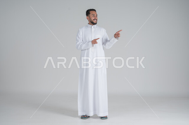 الوقوف باستقامة واعتدال, الإشارة إلى شيء ما, بورتريه لرجل عربي خليجي سعودي يرتدي الثوب التقليدي يشير بيديه إلى شيء ما, صورة الجسم بالطول الكامل, خلفية رمادي