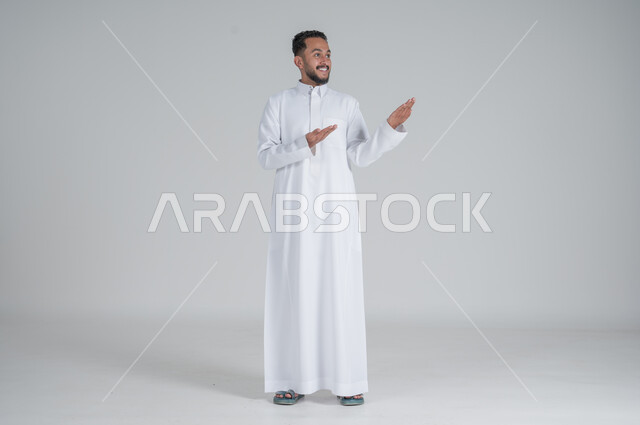 الوقوف باستقامة واعتدال, الإشارة إلى شيء ما, بورتريه لرجل عربي خليجي سعودي يرتدي الثوب التقليدي يشير بيديه إلى شيء ما, صورة الجسم بالطول الكامل, خلفية رمادي