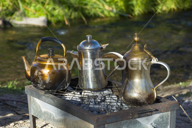 صورة لأباريق الشاي العربية الشرقية القديمة على كانون النار بخلفية من الطبيعة الخلابة بالسعودية ، التنزه والسفر ، صورة مقربة لأباريق الشاي المعدنية على موقد نار الفحم ، الطبيعة في السعودية