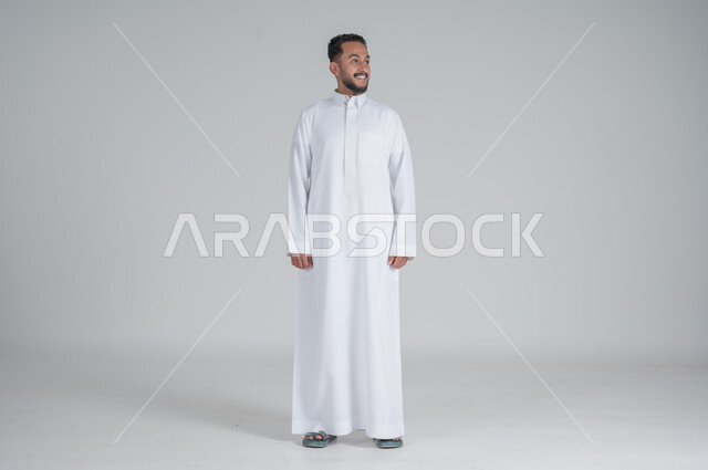 الوقوف باستقامة واعتدال, الشعور بالثقة في النفس, بورتريه لرجل عربي خليجي سعودي يرتدي الثوب التقليدي ينظر الى شيء ما, صورة الجسم بالطول الكامل, خلفية رمادي