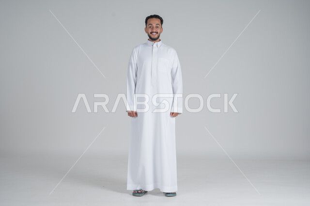 الوقوف باستقامة واعتدال, الشعور بالثقة في النفس, بورتريه لرجل عربي خليجي سعودي يرتدي الثوب التقليدي ينظر الى الكاميرا بتعابير السعادة والسرور, صورة الجسم بالطول الكامل, خلفية رمادي