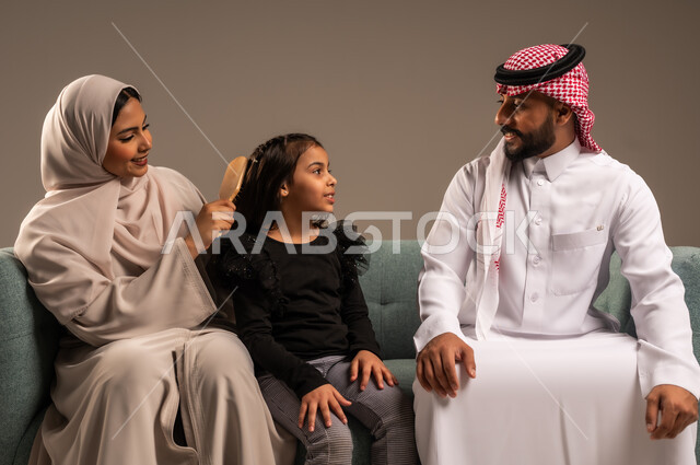مفهوم العناية والاهتمام بالأناقة والمظهر الخارجي، إيماءات السعادة والسرور، المحافظة على شعر صحي وأنيق، بورتريه لامراة عربية خليجية سعودية ترتدي عباءة ملونة تجلس على كرسي مريح مع زوجها وتقوم بتصفيف وتمشيط شعر ابنتها باستخدام الفرشاة ( المشط )، خلفية رمادية